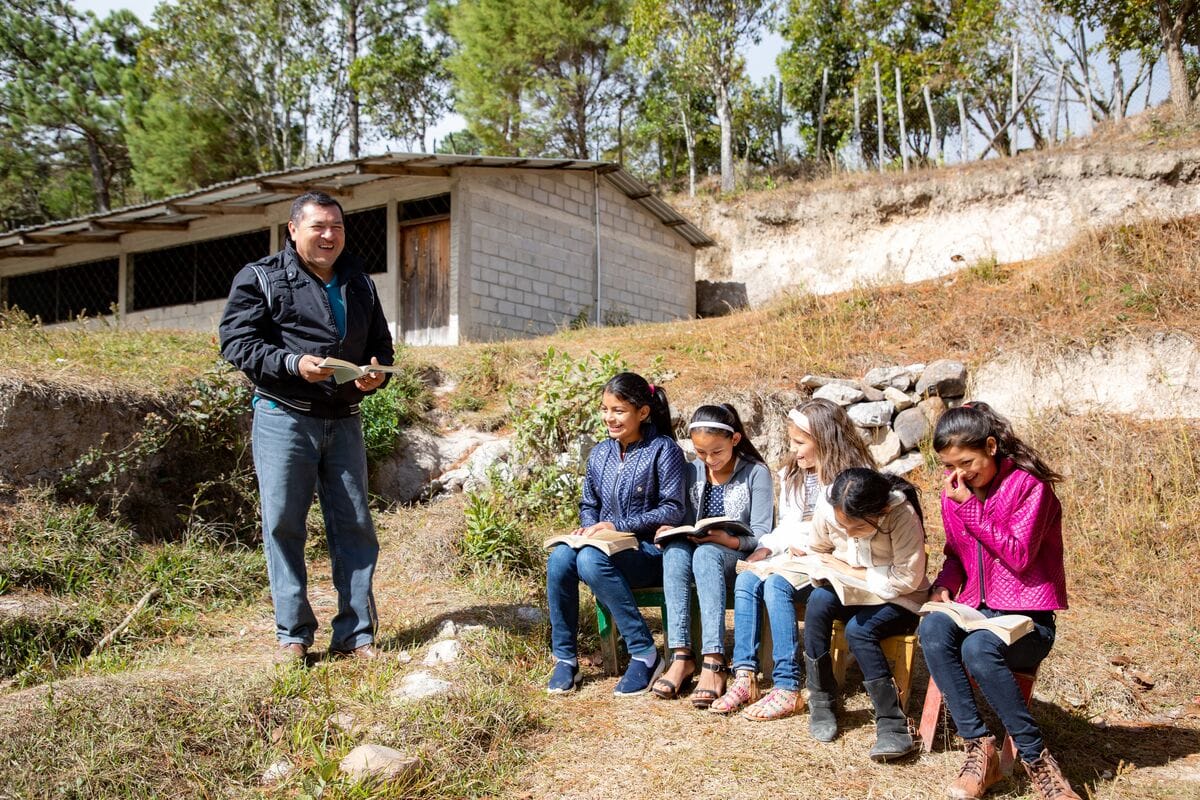 alt="Pastor Genaro in Honduras mit Patenkindern vor der Gemeinde. Kinder lesen Bibel"