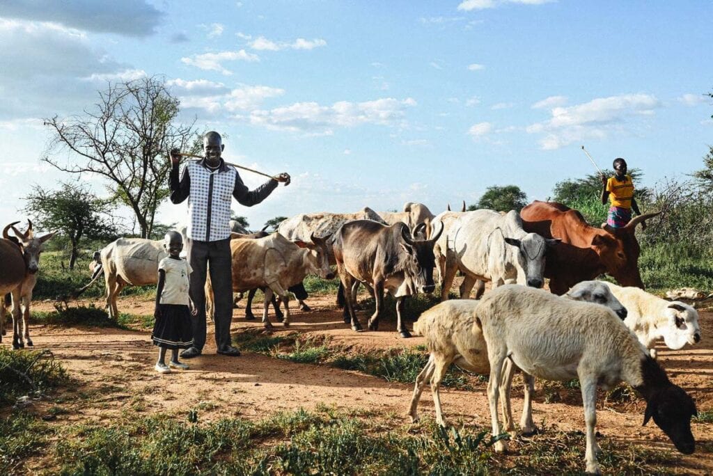 alt="Eli mit Tochter auf Feld mit Ziegen und Kühen Geschichte Uganda Lebensmittelknappheit Überleben Diebstahl Compassion Deutschland"