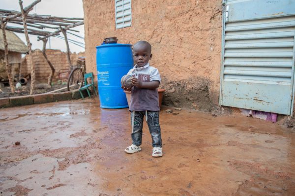 alt="Hunger kostet Würde, Ein Junge steht mit einem Ball im Matsch vor einem Haus, Lebensmittelkrise, Compassion Deutschland"