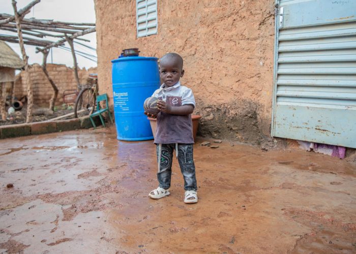 alt="Hunger kostet Würde, Ein Junge steht mit einem Ball im Matsch vor einem Haus, Lebensmittelkrise, Compassion Deutschland"