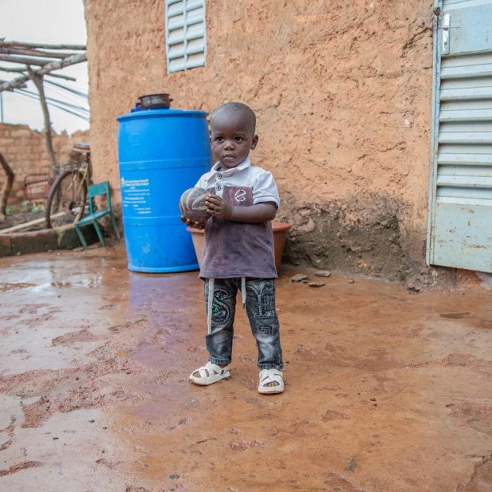 alt="Hunger kostet Würde, Ein Junge steht mit einem Ball im Matsch vor einem Haus, Lebensmittelkrise, Compassion Deutschland"